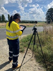 Rong standing near a road wearing high-vis and monitoring noise output.